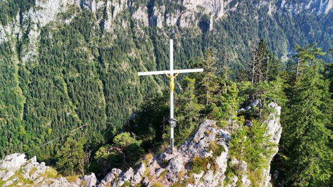 Witte kruis op een rots boven een groene bergvallei met bomen.