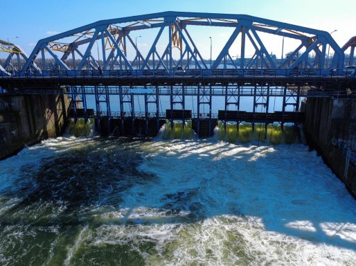 Een metalen brug boven een waterweg met stromend water en algen op de bodem.