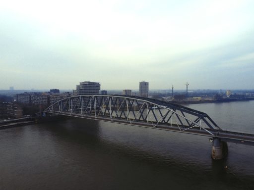 Stalen boogbrug over een rivier met gebouwen op de achtergrond en een bewolkte lucht.
